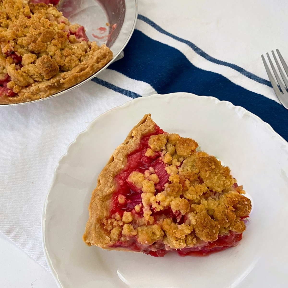 Overhead view of a slice of Strawberry Rhubarb Pie with Crumb Topping on a white plate, with the remaining pie and a fork in the background.