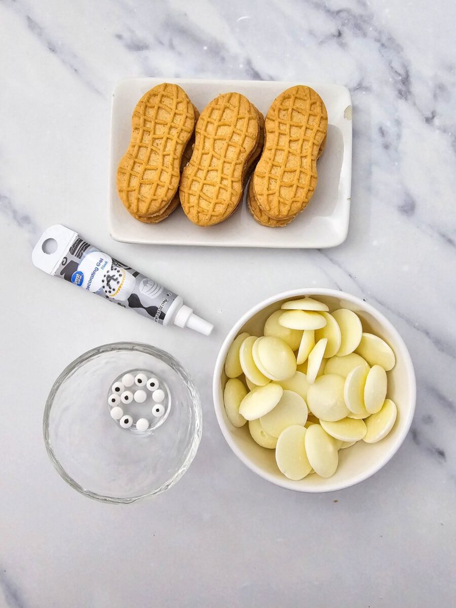 Nutter Butter cookies, white candy melts, candy eyeballs, and black decorating gel arranged on a white marble surface.