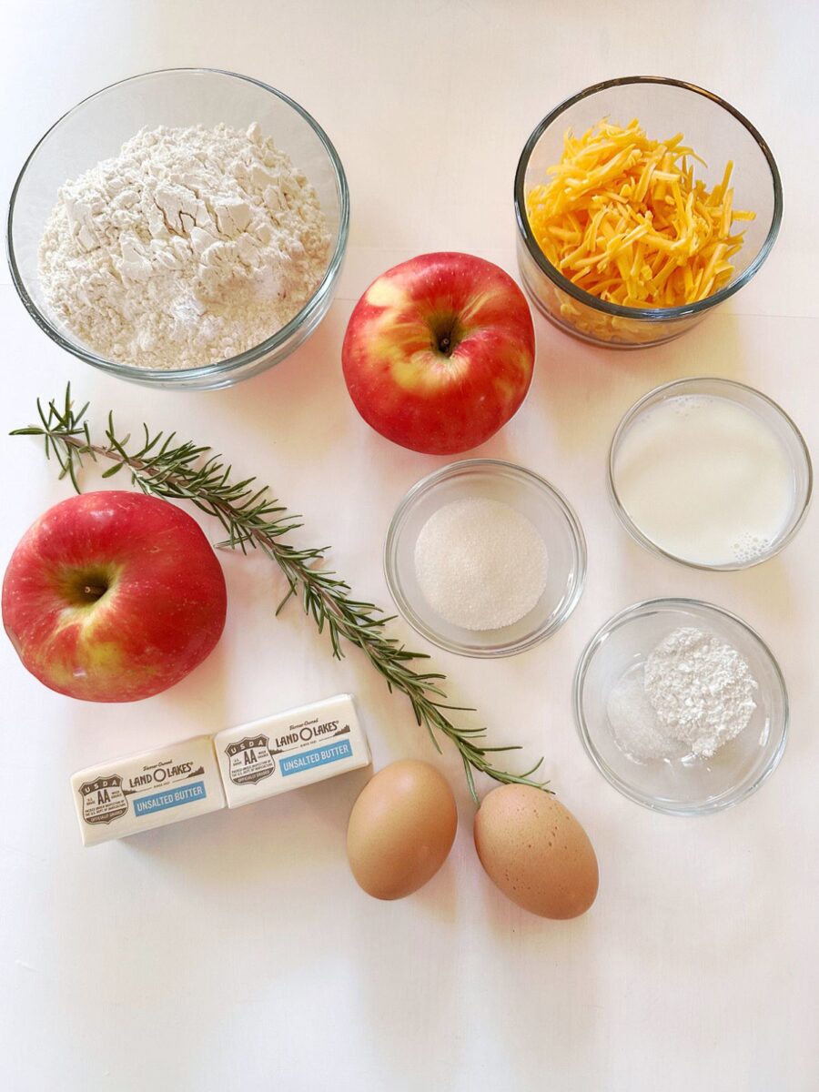 Apples, rosemary, flour, shredded cheddar, milk, sugar, baking powder, butter, and eggs arranged on a white surface.
