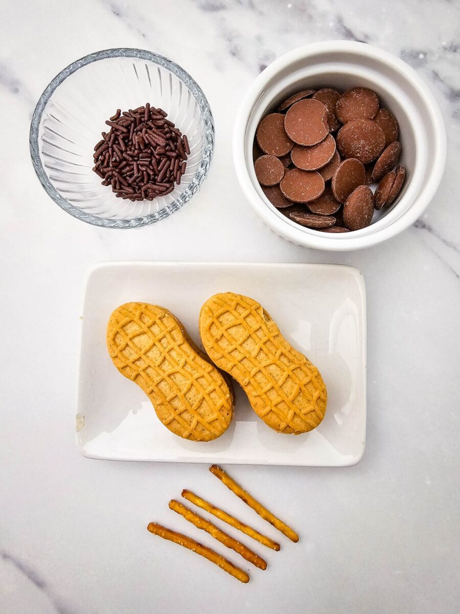 Nutter Butter cookies, chocolate melts, chocolate sprinkles, and pretzel sticks arranged on a white marble surface.
