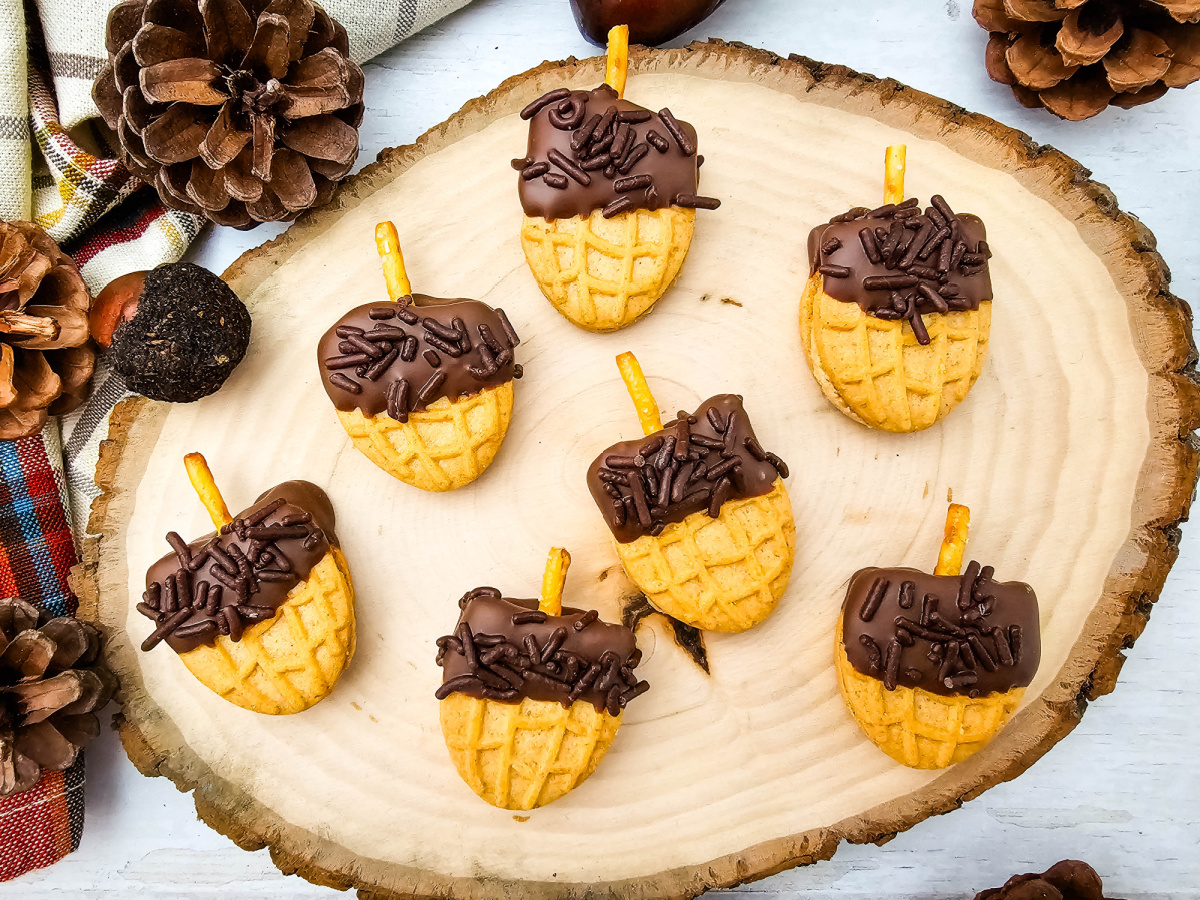 Seven acorn-shaped cookies arranged on a rustic wood slice with pinecones, plaid fabric, and decorative acorns.