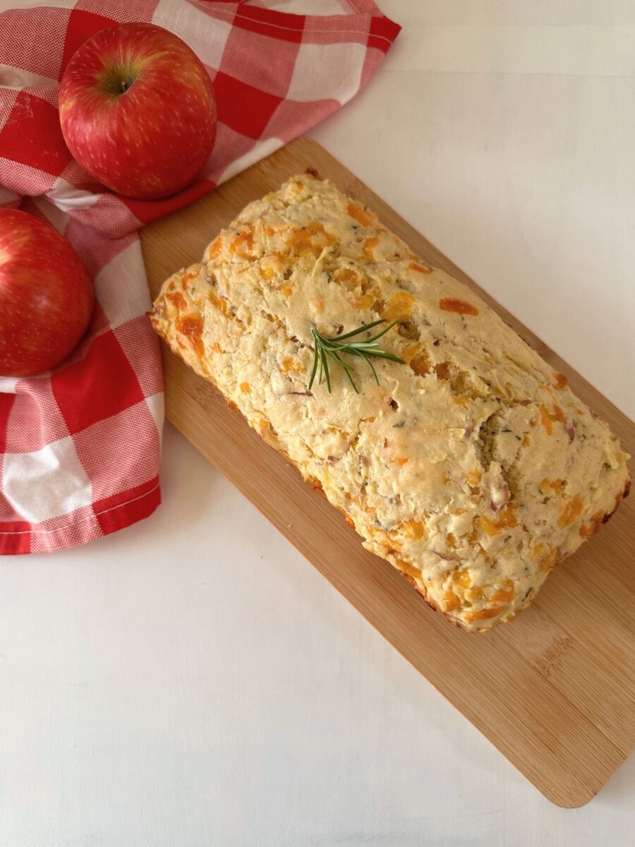 Whole savory apple and cheese loaf garnished with rosemary on a wooden cutting board beside two red apples and a checked cloth.