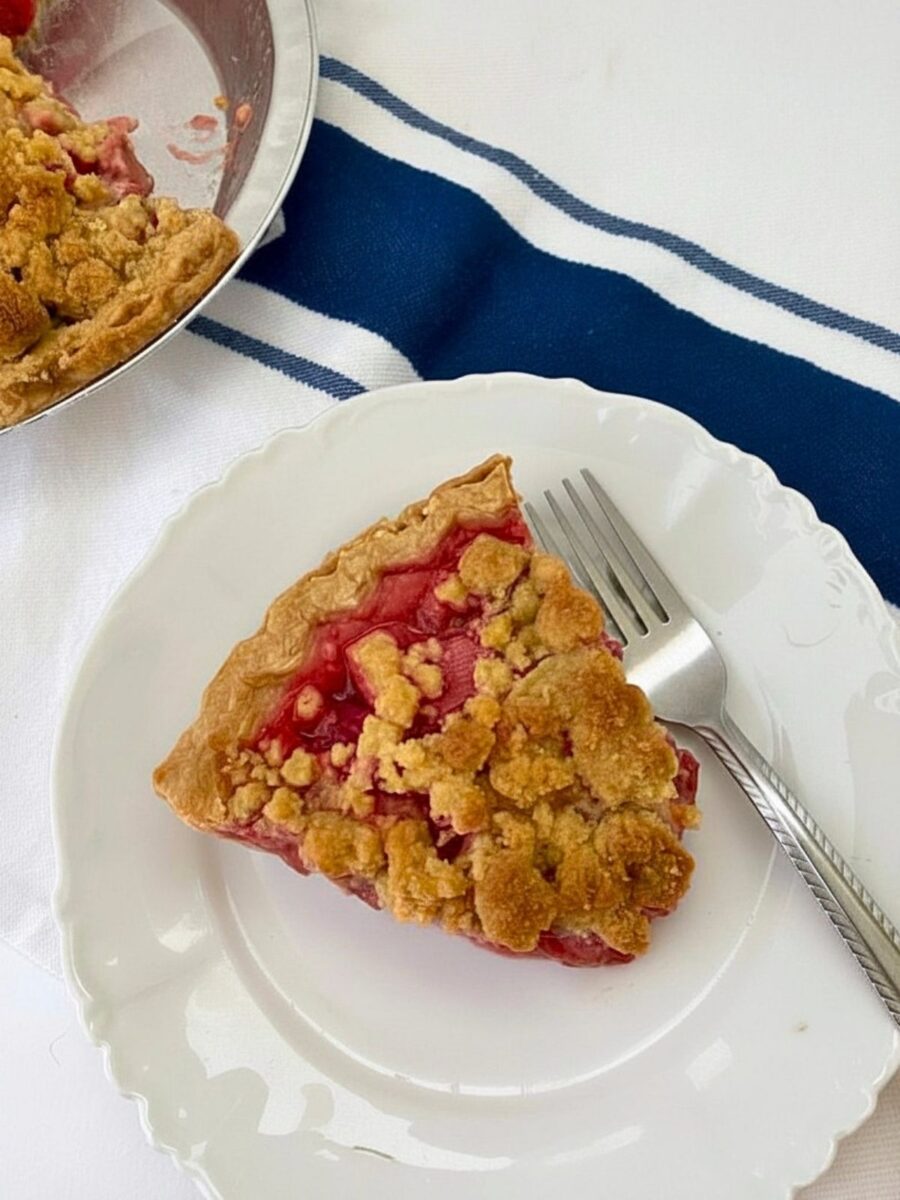 Overhead view of a strawberry rhubarb crumble pie slice on a floral white plate beside a fork. Part of the remaining pie is visible in the upper-left corner.