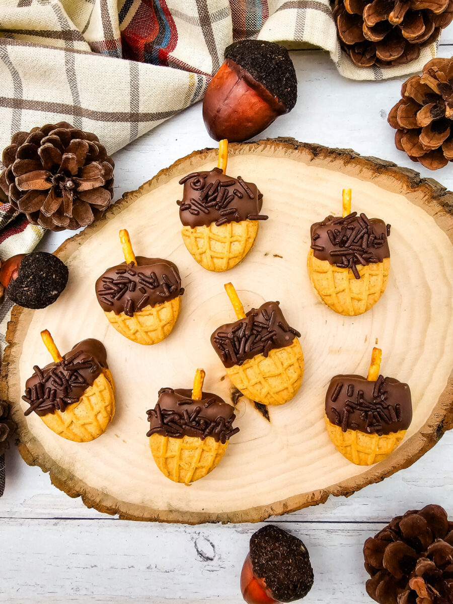 Overhead view of chocolate-dipped acorn cookies on a round wooden board surrounded by pinecones, plaid fabric, and fall decorations.