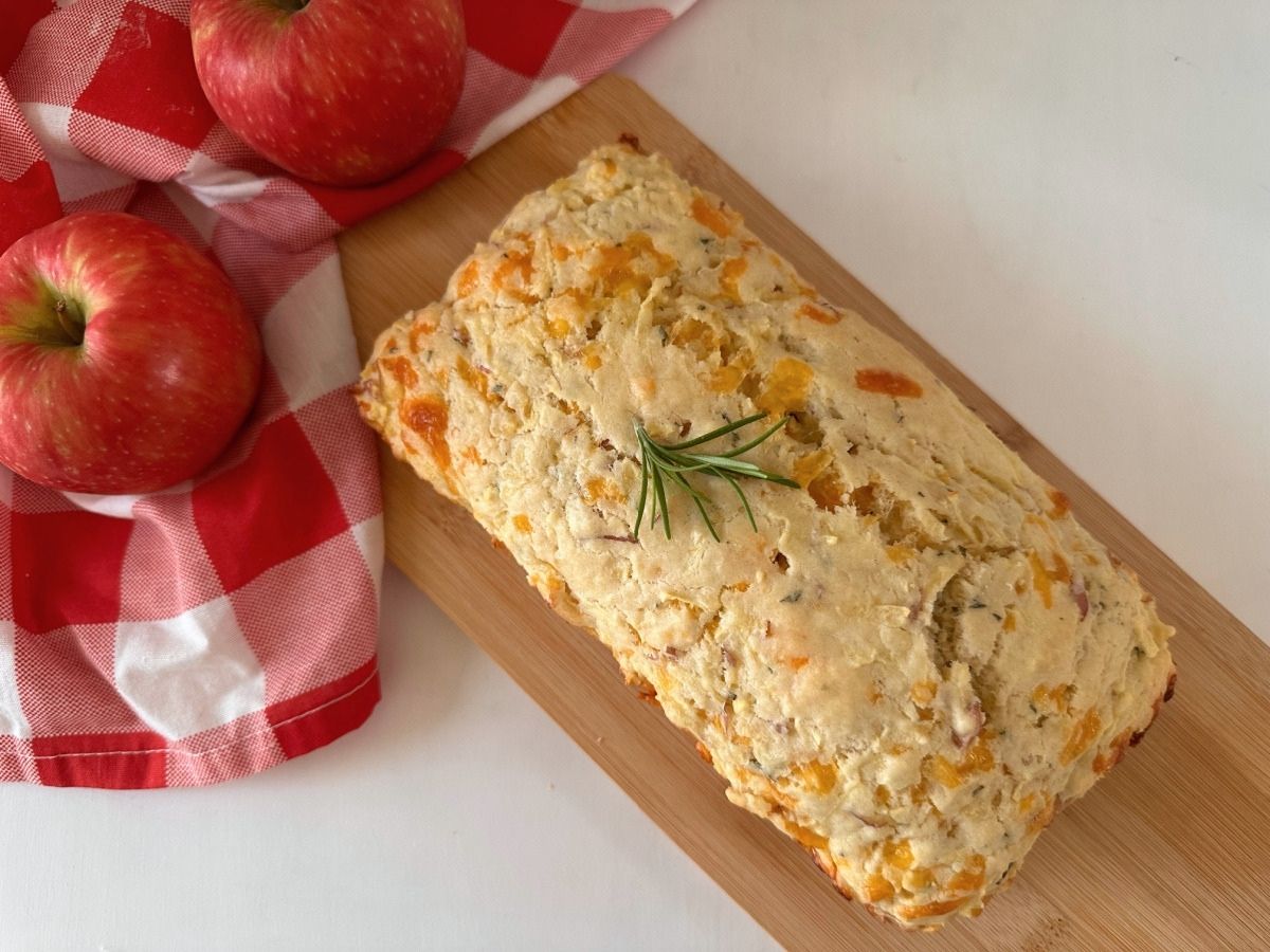 Overhead view of a whole baked loaf topped with rosemary, with red apples and a checked cloth beside the wooden cutting board.