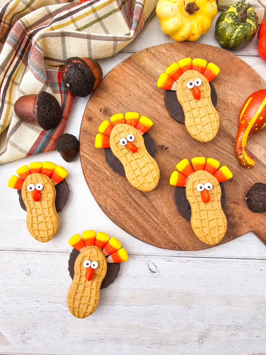 Five decorated Nutter Butter turkey cookies displayed around a wooden board with plaid fabric, acorns, and colorful fall gourds.