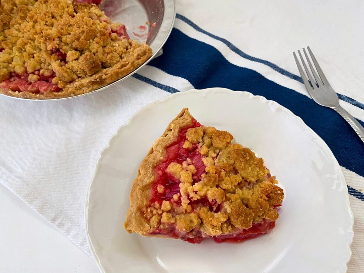 A plated slice of strawberry rhubarb pie with golden crumb topping, with the partially sliced pie in its baking pan and a fork in the background.