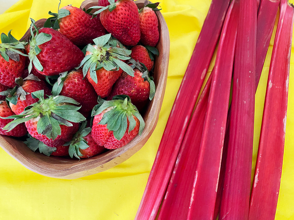 Fresh strawberries in a wooden bowl beside several bright red rhubarb stalks on a yellow background.