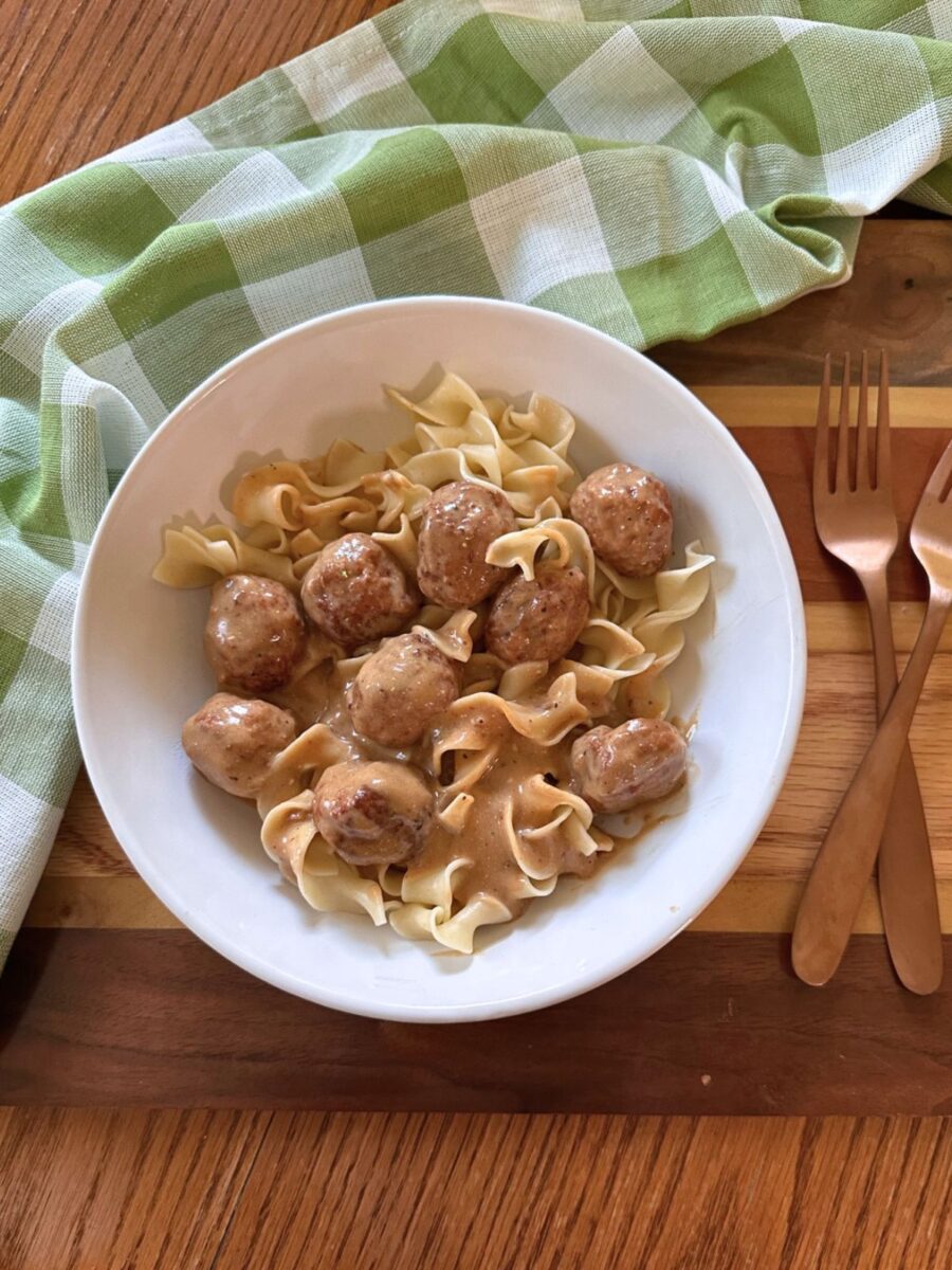 White bowl filled with creamy Swedish meatballs and egg noodles on a wooden tray with copper forks and a green gingham towel.