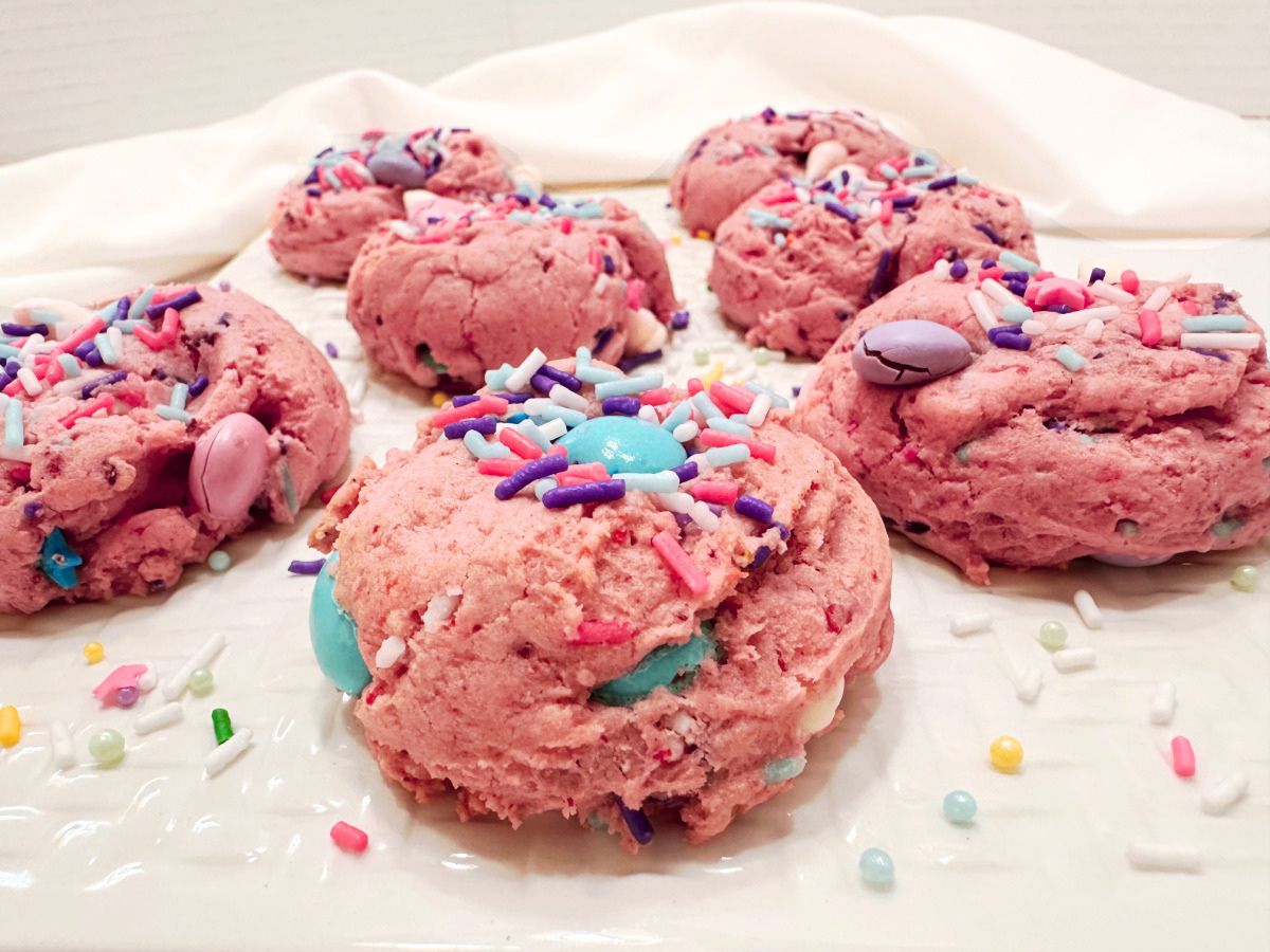 Close-up of thick pink strawberry cake mix cookies on a white platter, topped with blue, pink, and purple candy pieces, white baking chips, and unicorn sprinkles.