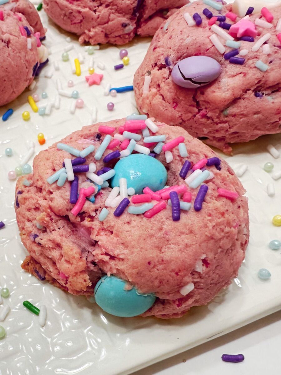 Close-up of a pink strawberry cookie topped with a turquoise candy piece and pink, purple, pale blue, and white sprinkles, with more cookies behind it.