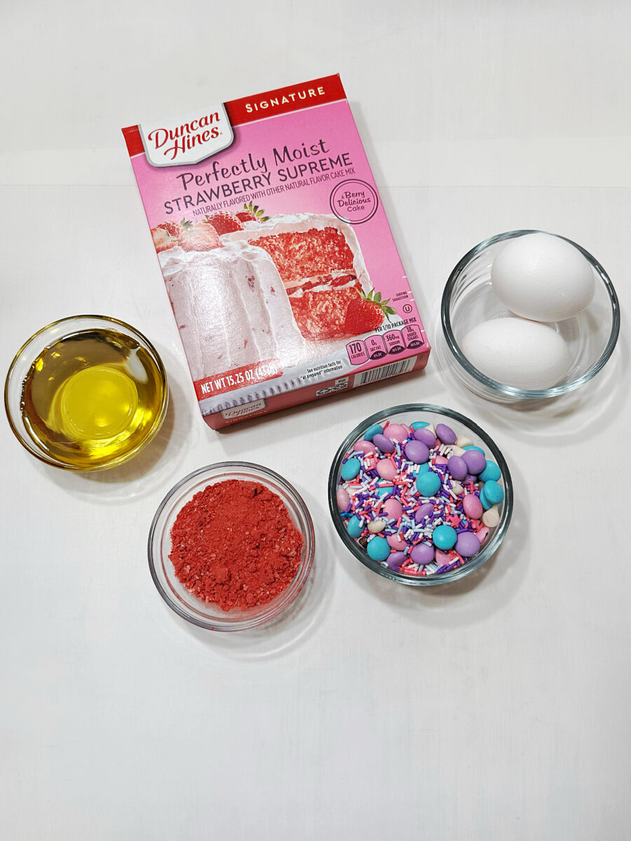 Strawberry cake mix beside glass bowls of eggs, avocado oil, finely crushed freeze-dried strawberries, and unicorn baking confetti on a white surface.